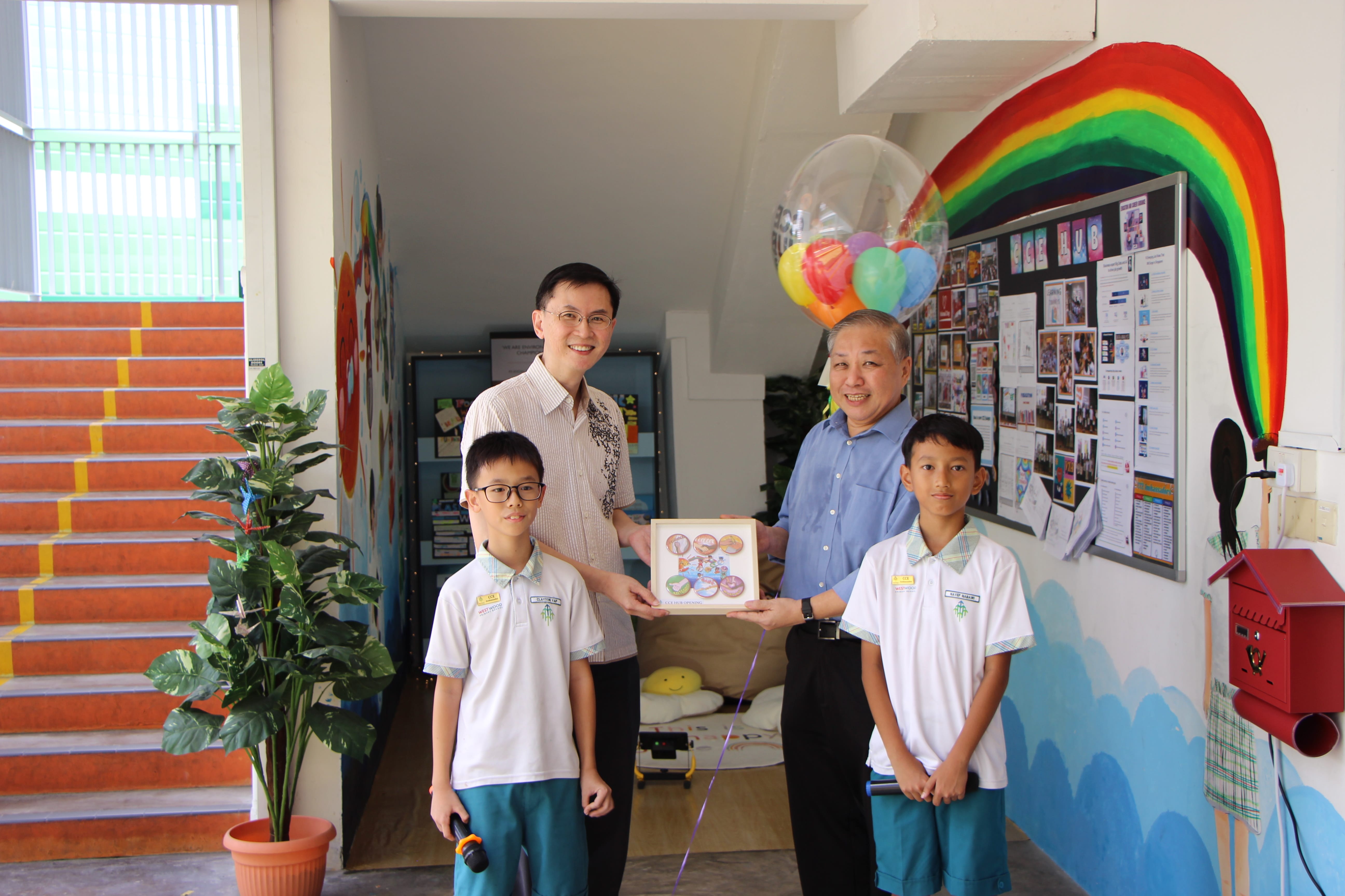 Two men and two schoolboys pose indoors holding a certificate, with balloons and a rainbow mural behind.